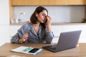 Eine junge Frau sitzt am Küchentisch vor einem Laptop und greift sich gestresst ins Gesicht. Sie braucht Stressmanagement Training.