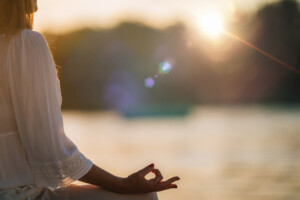 Eine Frau übt bei untergehender Sonne im Meditationssitz vor einem See präsent bleiben