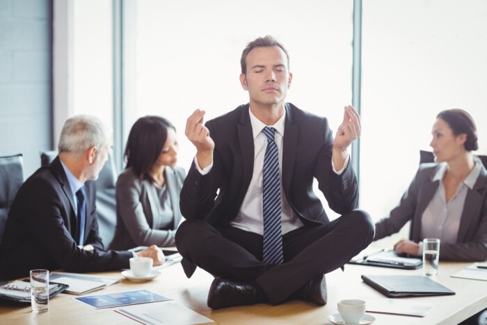 Ein Mann sitzt im Anzug mitten in einem Business Meeting meditierend auf einem Tisch, um gegen Angst und Depression vorzugehen