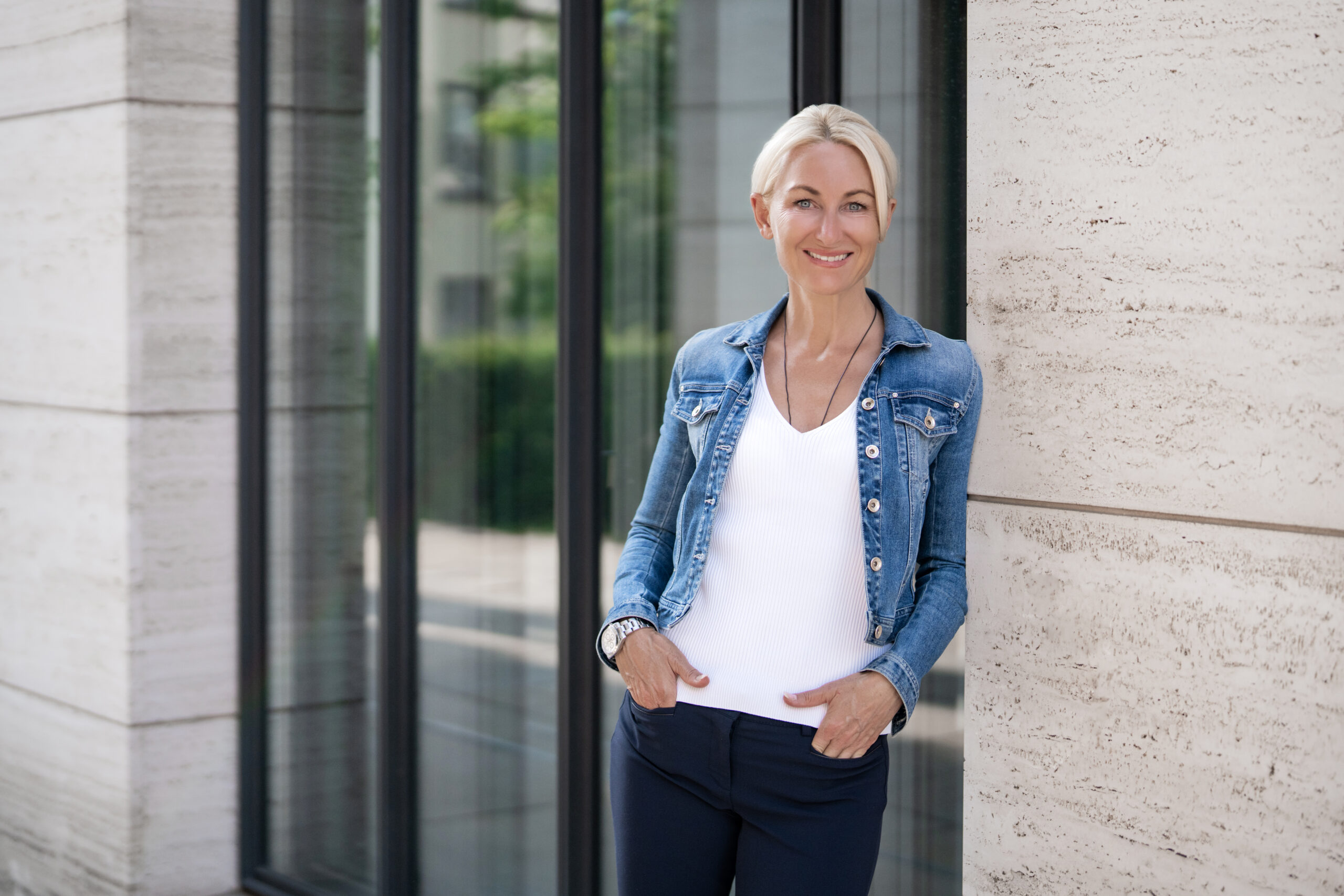 Kerstin Hardt lächelnd in Jeansjacke vor einem Fenster mit Sandsteinfassade zeigt, wie einfach mentale Stressbewältigung ist