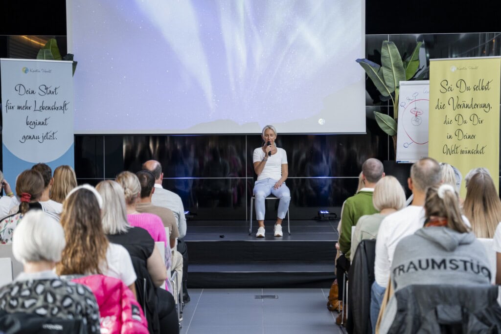 Kerstin Hardt auf einer Bühne vor einer Menschenmenge mit einem Mikrophon in der Hand leitet eine Meditation zum endlich wieder glücklich sein 