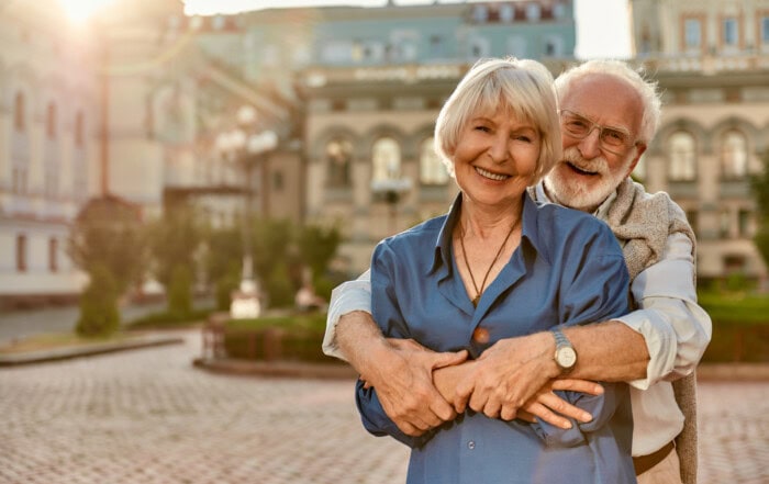 Ein Pärchen steht, der Mann umarmt die Frau von hinten, vor einem Gebäude. Die beiden lachen und zeigen so, dass man auch im Alter jung bleiben kann.